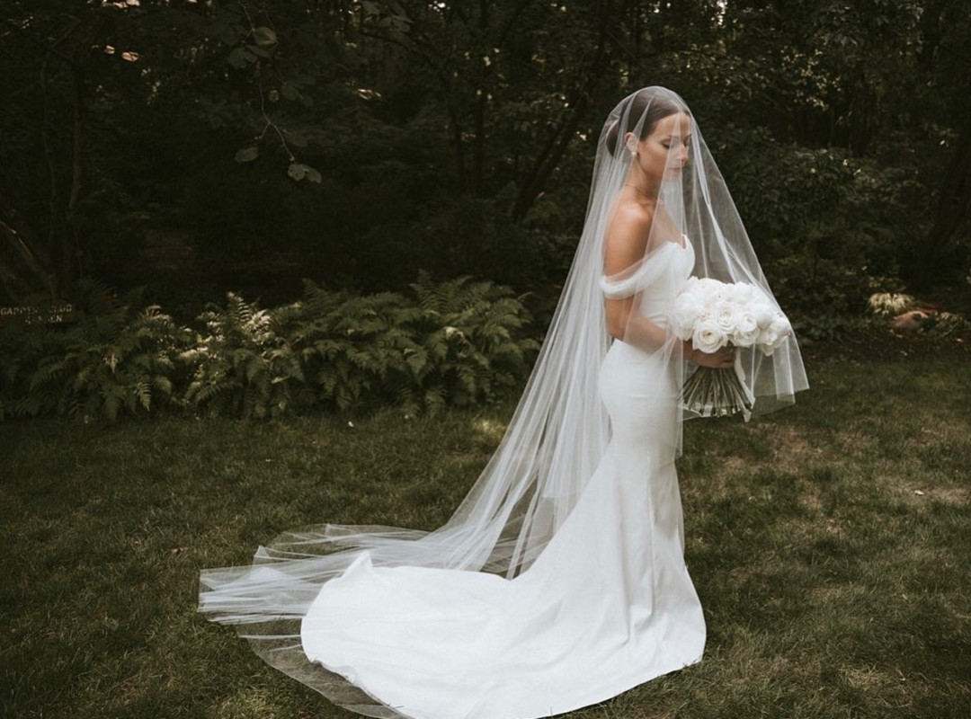 woman wearing a wedding dress and a wedding veil with a blusher holding white flowers in an outdoor garden