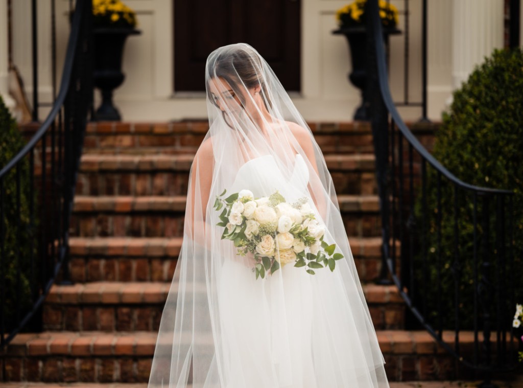 Woman wearing a bridal veil with a long blusher in front of her face and her flowers