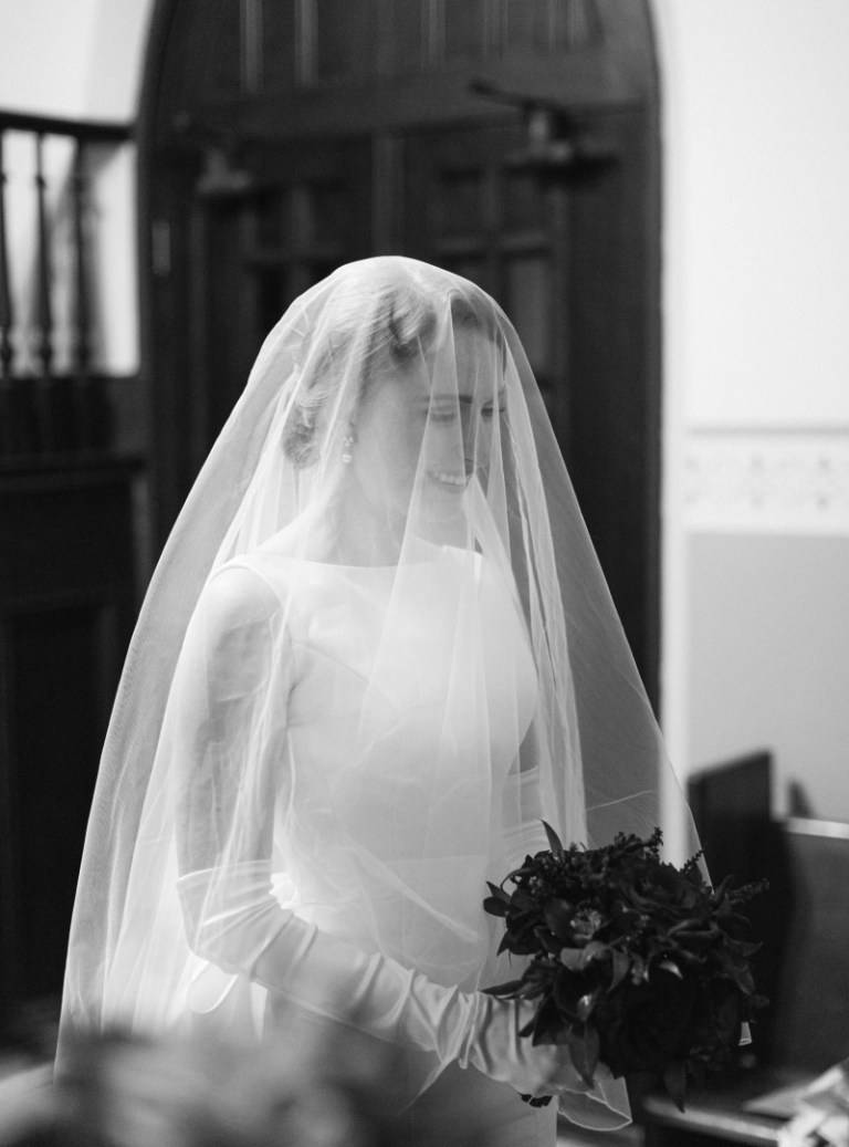 black and white photo of a woman wearing a wedding gown with a bridal veil in front of her face against a dark background