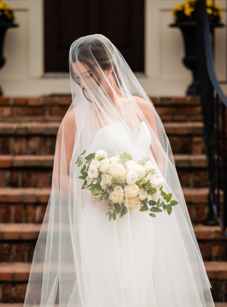 Photo of a woman wearing a wedding dress with a bridal veil over her face in front of an outdoor staircase